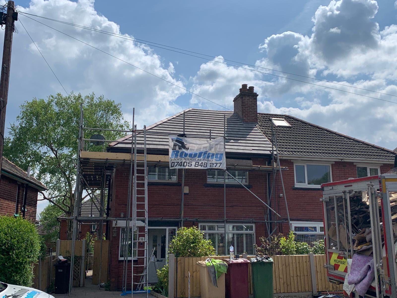 A red brick semi-detached house with scaffolding up and a UK Rhino Roofing banner reading Roofs tough as Rhino, with the phone number